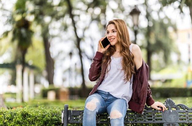 Giovane ragazza sorridente con il suo smartphone seduta su una panchina nel formato parco con spazio copia da vicino