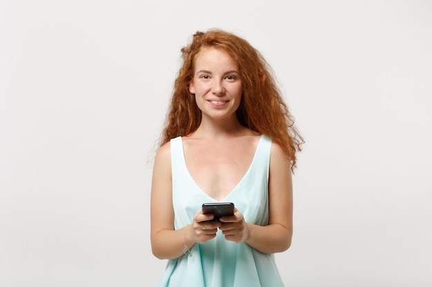 Giovane ragazza sorridente della donna della testarossa in vestiti leggeri casuali che posano isolati su fondo bianco, ritratto dello studio. Concetto di stile di vita della gente. Mock up copia spazio. Utilizzando il telefono cellulare, digitando un messaggio sms.