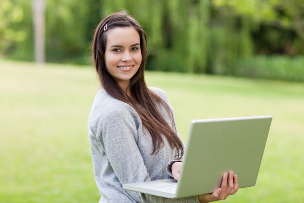 Giovane ragazza sorridente in piedi in un giardino pubblico mentre si utilizza il suo computer portatile