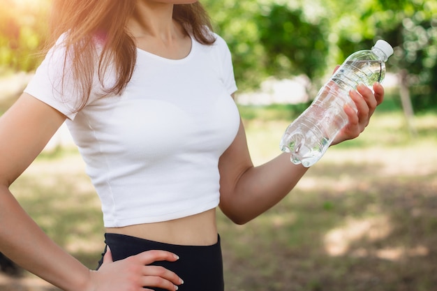 Giovane ragazza sottile in una maglietta bianca che tiene una bottiglia di acqua minerale.