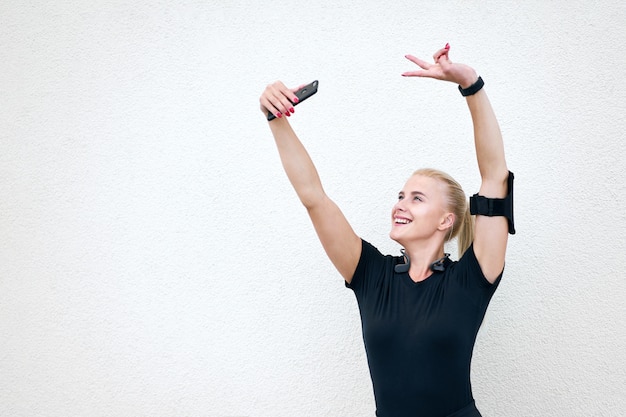 Giovane ragazza sportiva attraente che indossa abiti sportivi neri che ascolta la musica e che allunga sul fondo bianco della parete