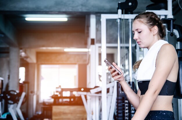 Giovane ragazza sportiva che riposa dopo un allenamento intenso in palestra con smartphone ascoltando musica