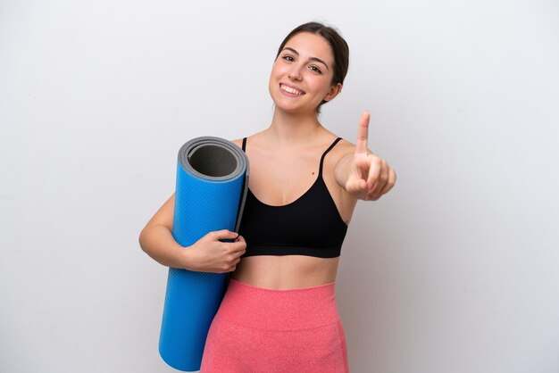 Giovane ragazza sportiva che va a lezioni di yoga mentre si tiene un tappetino isolato su sfondo bianco che mostra e solleva un dito