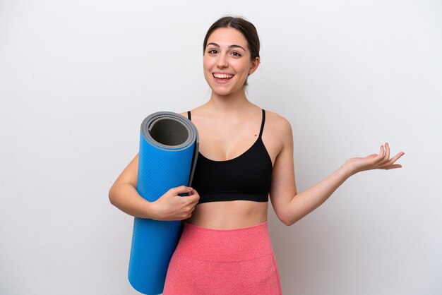 Giovane ragazza sportiva che va a lezioni di yoga mentre tiene un tappetino isolato su sfondo bianco con espressione facciale scioccata