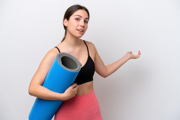 Giovane ragazza sportiva che va a lezioni di yoga mentre tiene un tappetino isolato su sfondo bianco estendendo le mani di lato per invitare a venire