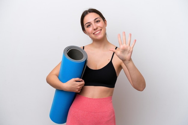 Giovane ragazza sportiva che va a lezioni di yoga tenendo un tappetino isolato su sfondo bianco contando cinque con le dita