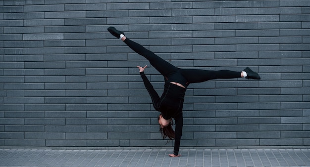 Giovane ragazza sportiva in abbigliamento sportivo nero che fa esercizi di verticale duri all'aperto vicino al muro grigio