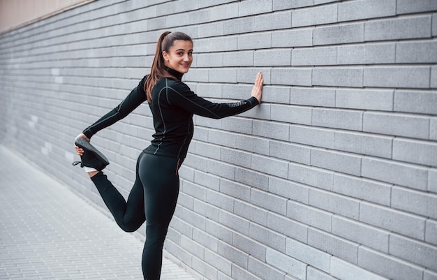 Giovane ragazza sportiva in abiti sportivi neri all'aperto facendo stretching vicino al muro grigio