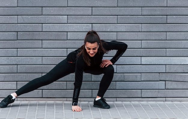 Giovane ragazza sportiva in abiti sportivi neri all'aperto facendo stretching vicino al muro grigio