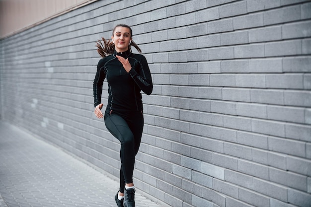 Giovane ragazza sportiva in abiti sportivi neri che corre all'aperto vicino al muro grigio