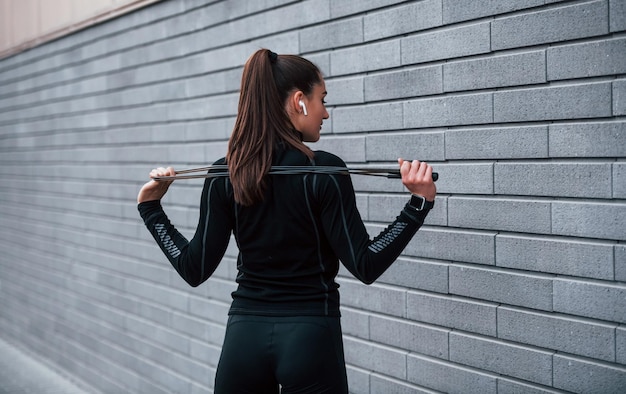 Giovane ragazza sportiva in abiti sportivi neri in piedi con la corda per saltare in mano all'aperto vicino al muro grigio