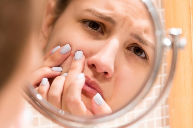 Giovane ragazza stringe il brufolo sul viso di fer davanti a uno specchio del bagno Concetto di bellezza per la cura della pelle e del benessere mattutino