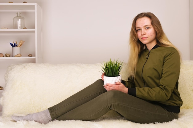 Giovane ragazza teenager con il fronte dell'acne che si siede sul sofà e che tiene l'immagine verde della pianta d'appartamento