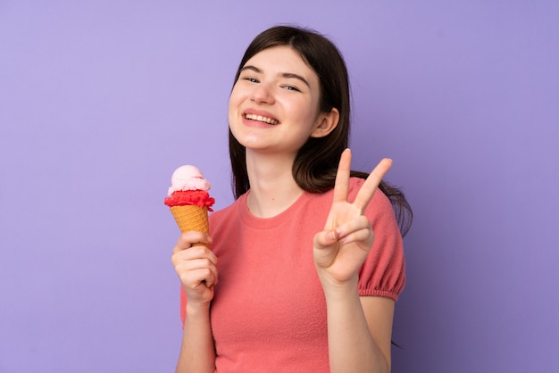 Giovane ragazza ucraina dell'adolescente che tiene un gelato della cornetta sopra la parete porpora che sorride e che mostra il segno di vittoria