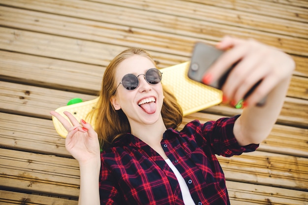 Giovane ragazza urbana alla moda in abito hipster che fa selfie mentre giaceva con sul molo di legno.