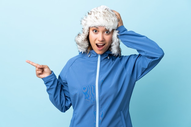 Giovane ragazza uruguaiana con cappello invernale isolato su blu sorpreso e puntando il dito verso il lato