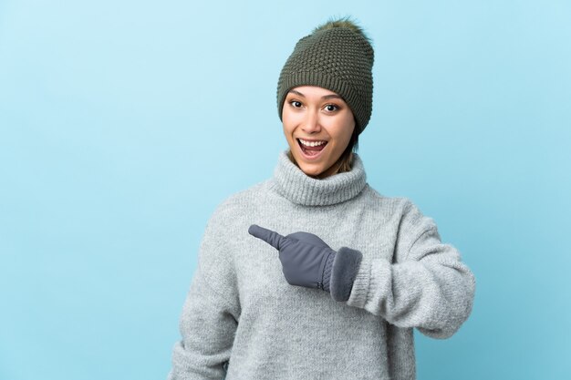 Giovane ragazza uruguaiana con cappello invernale sulla parete blu che punta il dito verso il lato