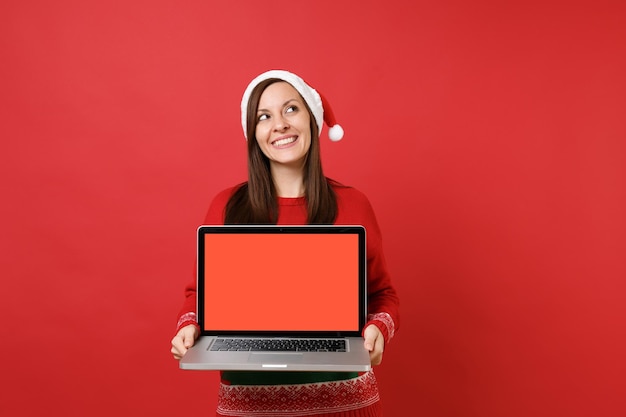 Giovane ragazza vaga di Santa che cerca tenendo il computer portatile del pc con lo schermo vuoto in bianco isolato sul fondo rosso luminoso della parete. Felice Anno Nuovo 2019 celebrazione festa concetto. Mock up copia spazio.