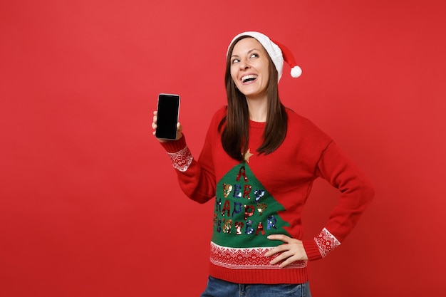 Giovane ragazza vaga di Santa in cappello di Natale che guarda in su, tenendo il telefono cellulare con schermo vuoto vuoto isolato su sfondo rosso. Felice Anno Nuovo 2019 celebrazione festa concetto. Mock up copia spazio.