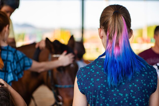 Giovane ragazza vista di spalle con i capelli colorati che guarda il cavallo e il braccio del caregiver