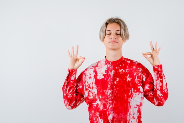 Giovane ragazzo adolescente in camicia che mostra il segno di mudra, tenendo gli occhi chiusi e guardando rilassato, vista frontale.