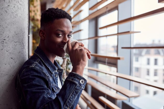 Giovane ragazzo afroamericano positivo in piedi al chiuso e bere caffè.