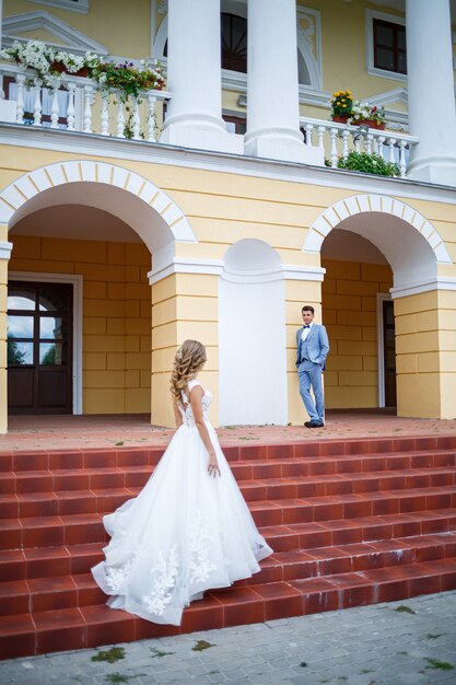 Giovane ragazzo alla moda nel costume dello sposo e della sposa bella ragazza in un vestito bianco con un treno che cammina sullo sfondo di una grande casa con colonne il giorno delle nozze