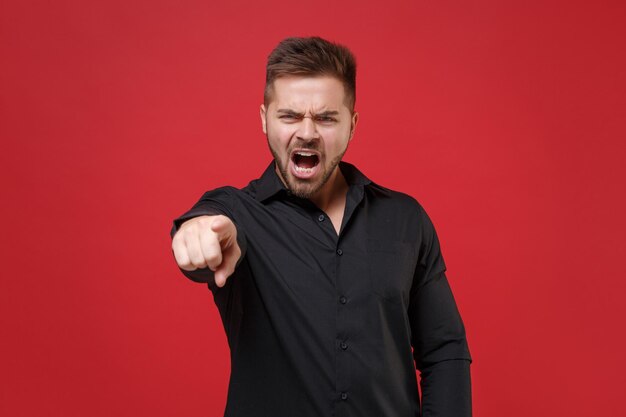 Giovane ragazzo barbuto irritato arrabbiato 20s in classica camicia nera in posa isolata su sfondo rosso studio. Concetto di stile di vita delle persone. Mock up spazio di copia. Puntare il dito indice sulla fotocamera urlando parolacce.