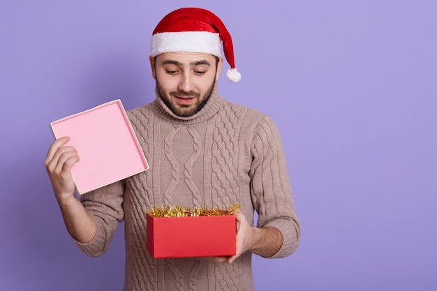 Giovane ragazzo barbuto sorpreso in maglione beige casual che apre il contenitore di regalo rosso