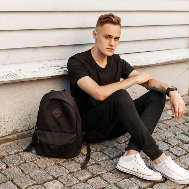 Giovane ragazzo bello in un vestito nero alla moda e scarpe bianche che si siede vicino alla parete di legno.