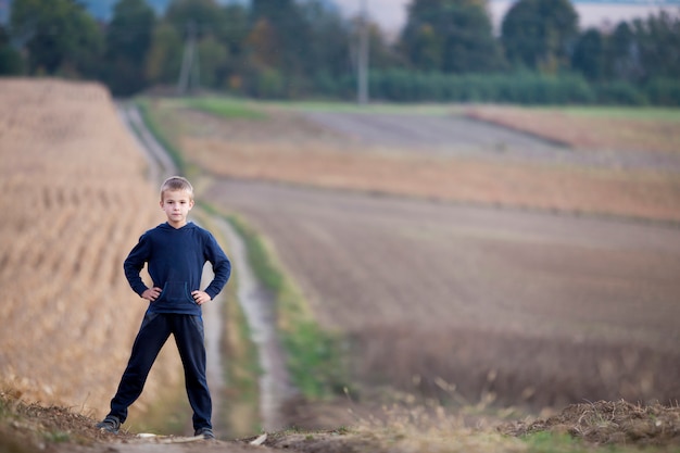 Giovane ragazzo biondo bello del bambino che sta da solo sulla strada a terra fra i campi di grano erbosi dorati sugli alberi verdi nebbiosi vaghi e sul fondo dei prati. Stile di vita attivo, concetto di attività del fine settimana esterno.