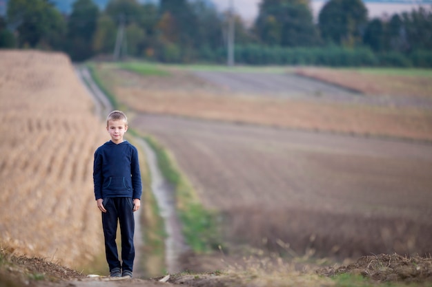 Giovane ragazzo biondo bello del bambino che sta da solo sulla strada a terra fra i campi di grano erbosi dorati sugli alberi verdi nebbiosi vaghi e sul fondo dei prati. Stile di vita attivo, concetto di attività di fine settimana all'aperto.