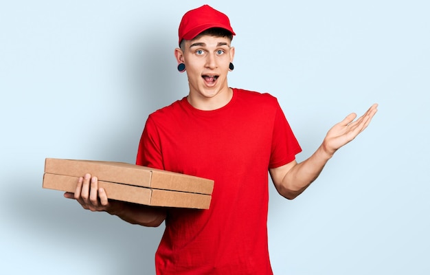 Giovane ragazzo caucasico con dilatazione delle orecchie che tiene la scatola della pizza di consegna che celebra il successo con un sorriso felice e l'espressione del vincitore con la mano alzata