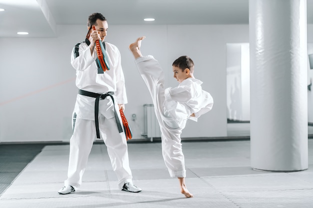 Giovane ragazzo caucasico in dobok calci a piedi nudi mentre trainer tenendo il bersaglio di calcio. Taekwondo concetto di formazione.