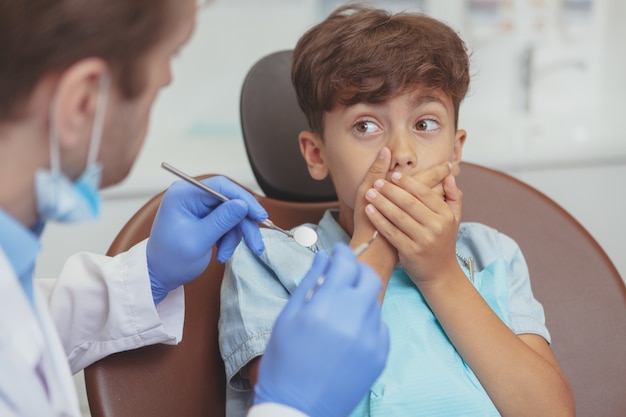 Giovane ragazzo che nasconde la bocca dietro le mani, rifiutando di ottenere il controllo dei denti dal dentista professionista