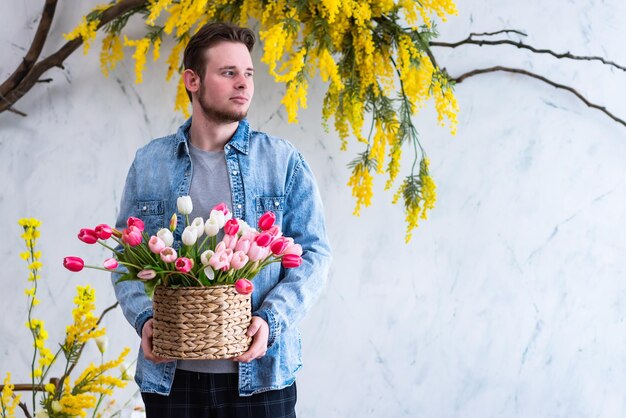 Giovane ragazzo con un cesto di vimini di tulipani colorati nelle sue mani posa contro un muro bianco testurizzato decorato con mimosa gialla