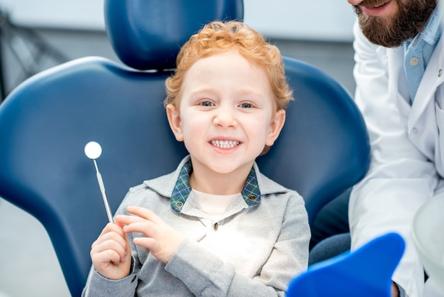 Giovane ragazzo eccitato che guarda lo specchio dentale seduto sulla sedia dello studio dentistico