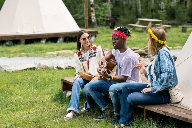 Giovane ragazzo in bandana seduto con le ragazze in tenda e cantando bella canzone al campeggio