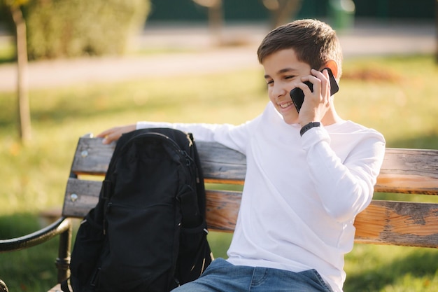 Giovane ragazzo in felpa bianca con zaino nero seduto sulla panchina nel parco e parla con qualcuno al telefono