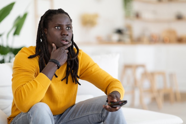 Giovane ragazzo nero stupito che tiene il telecomando e guarda lo schermo della TV