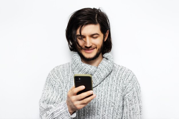 giovane ragazzo sorridente con lo smartphone in mano. Indossare maglione a collo alto sul muro bianco.