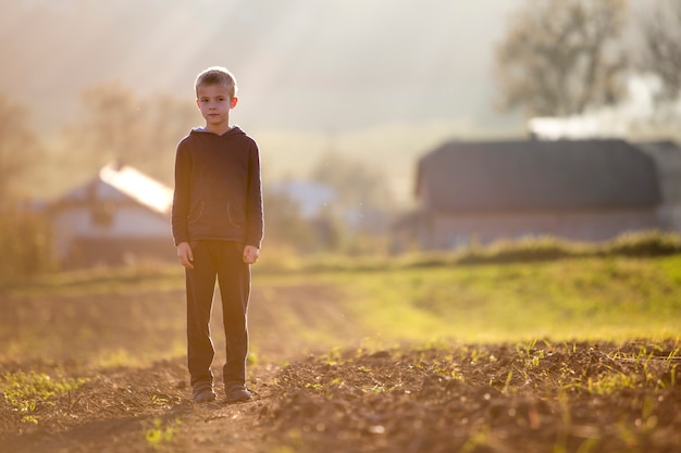 Giovane ragazzo stanco serio biondo del bambino che sta da solo sul campo dopo il raccolto il giorno soleggiato di fine dell'estate o di autunno.