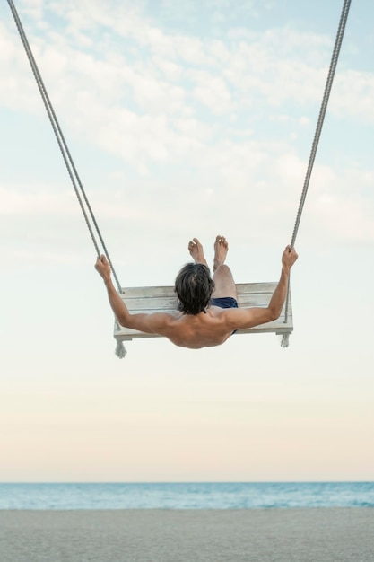 Giovane ragazzo sull'altalena della corda sulla spiaggia sullo sfondo del mare Cornice verticale Riposo sul mare Vista posteriore