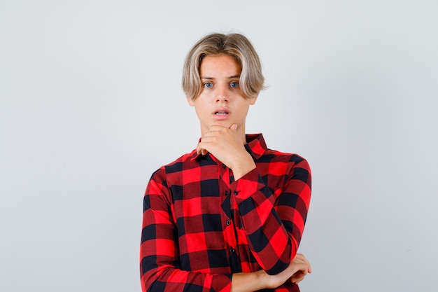 Giovane ragazzo teenager che tiene la mano sul mento in camicia a quadri e sembra sorpreso. vista frontale.