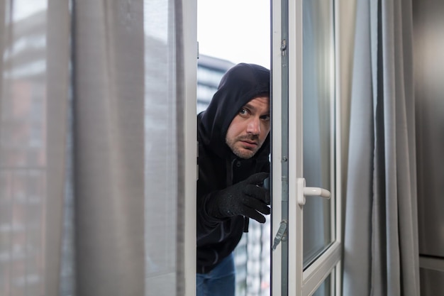 Giovane rapinatore maschio in abiti scuri guarda fuori dalla finestra del condominio Vuole andare in un alloggio