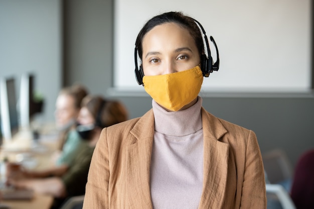 Giovane rappresentante dell'assistenza clienti femminile di razza mista in cuffia, maschera protettiva e abbigliamento casual in piedi contro la fila di colleghi