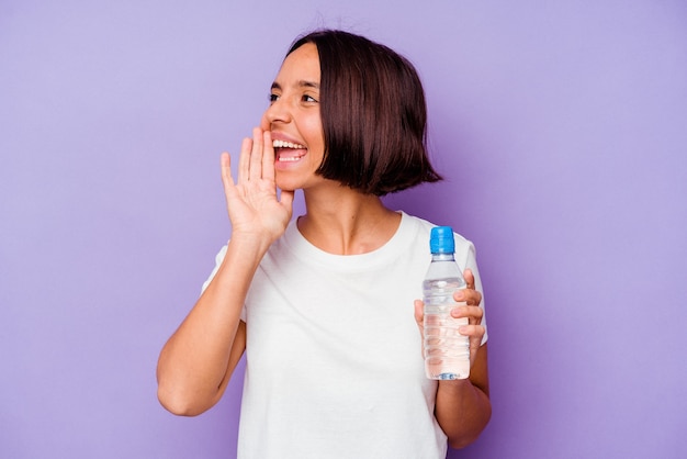 Giovane razza mista che tiene una bottiglia d'acqua isolata su sfondo viola gridando e tenendo il palmo vicino alla bocca aperta.
