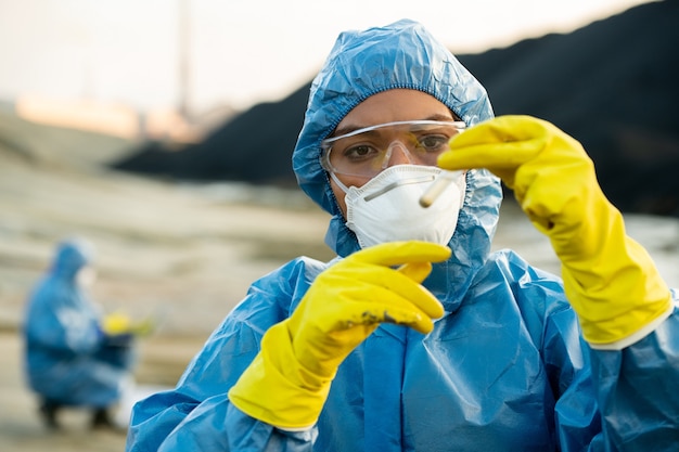 Giovane ricercatrice in abiti da lavoro protettivi che tiene un campione di terreno tossico in un pallone davanti al viso durante un'indagine ecologica con un collega