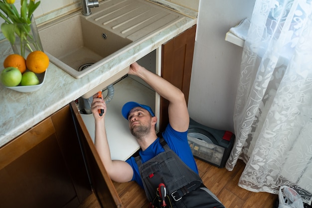 Giovane riparatore con strumenti in mano e un berretto blu sta riparando il lavandino in cucina