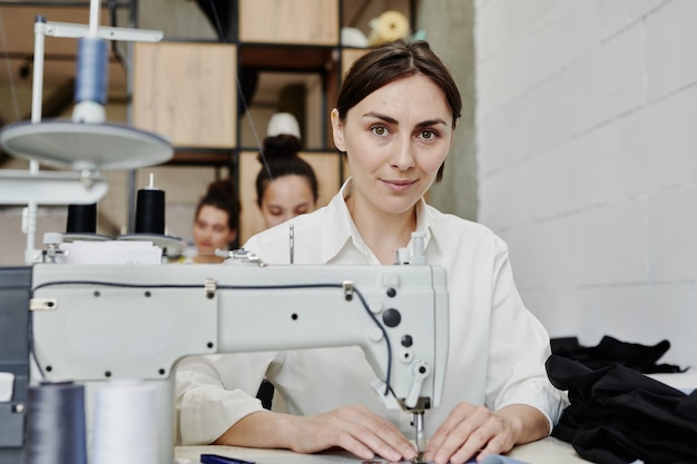 Giovane sarta contemporanea ti guarda mentre è seduto sul posto di lavoro con la macchina da cucire elettrica e fa o ripara vestiti